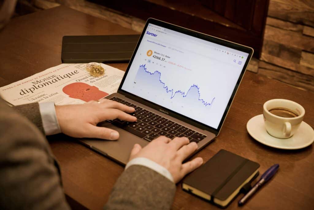 A person in a suit uses a laptop displaying a fluctuating stock chart, possibly reviewing fixed asset management software. On the table are a newspaper, cup of coffee, notebook, pen, and a small globe.