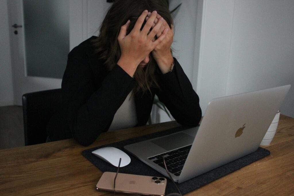 A person sits at a desk with their head in their hands, appearing stressed in front of an open laptop—perhaps struggling with double bookings while managing their rental business without rental reservation software. A smartphone, eyeglasses, and mouse are on the desk.