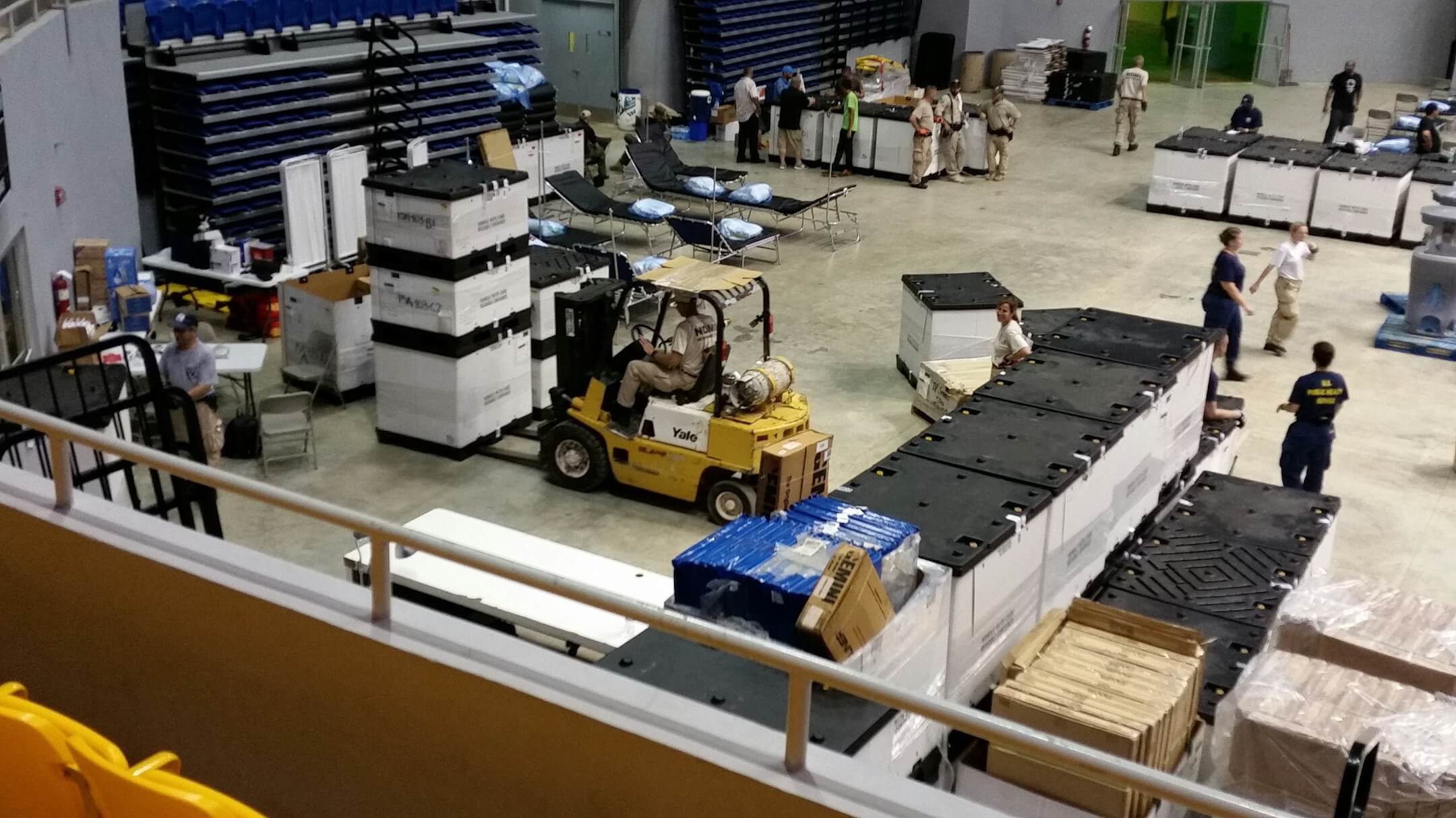 A forklift moves large black and white containers in a gymnasium where people in uniforms, aided by rental reservation software, organize supplies and cots, likely for emergency shelter or disaster relief.
