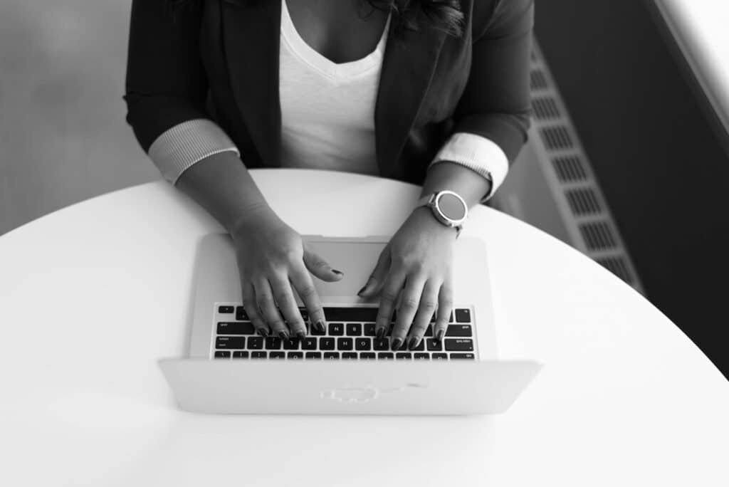Black and white image of a person typing on a laptop at a round white table, possibly working with fixed asset management software. The person wears a blazer, white shirt, and smartwatch; the overhead view focuses on their hands.