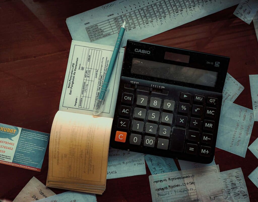 A black Casio calculator, blue pen, receipts, handwritten notes, and a business card are spread out on a wooden desk—suggesting a rental business owner managing financials or avoiding double bookings with the help of rental reservation software.