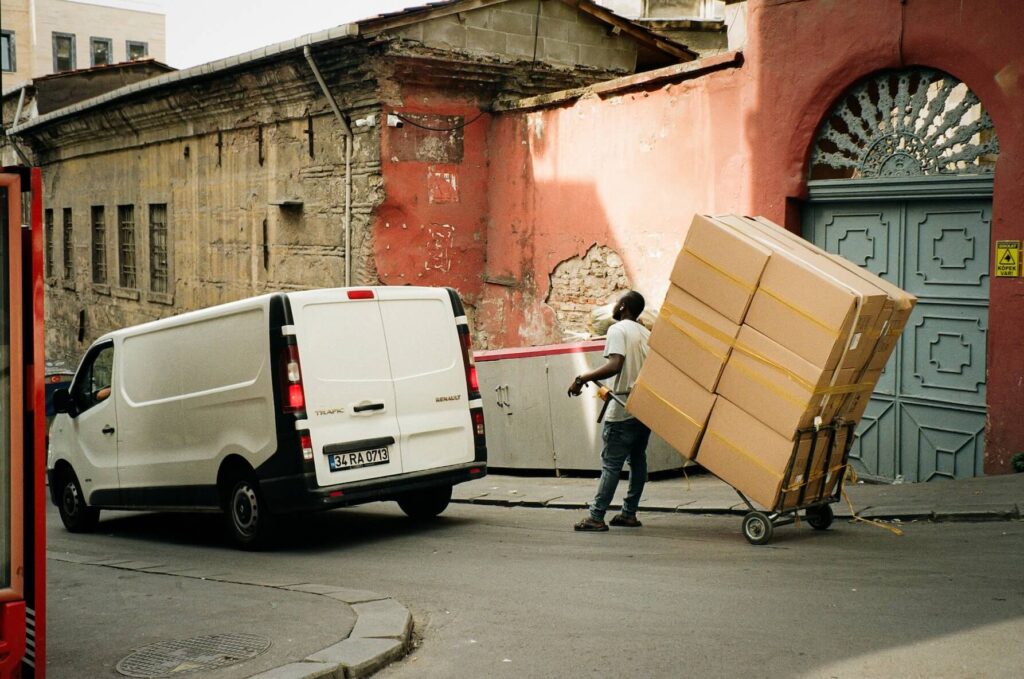 A man pushes a loaded cart with large cardboard boxes up a street next to a white van and an old, weathered building, perhaps making deliveries for a rental business with reliable rental reservation software.
