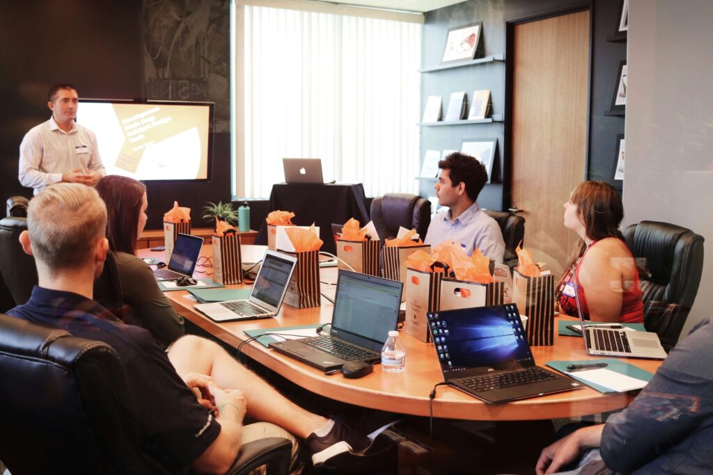 A man gives a presentation on fixed asset management software to five seated people in a modern conference room, with laptops and gift bags on the table and a large screen displaying a slide in the background.