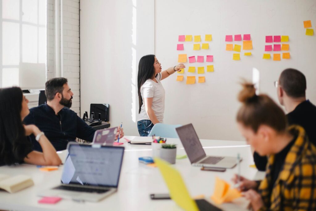 A woman stands at a whiteboard covered with colorful sticky notes, leading a discussion on improving their rental business and preventing double bookings with four colleagues seated at a table with laptops and notebooks in a bright, modern office.