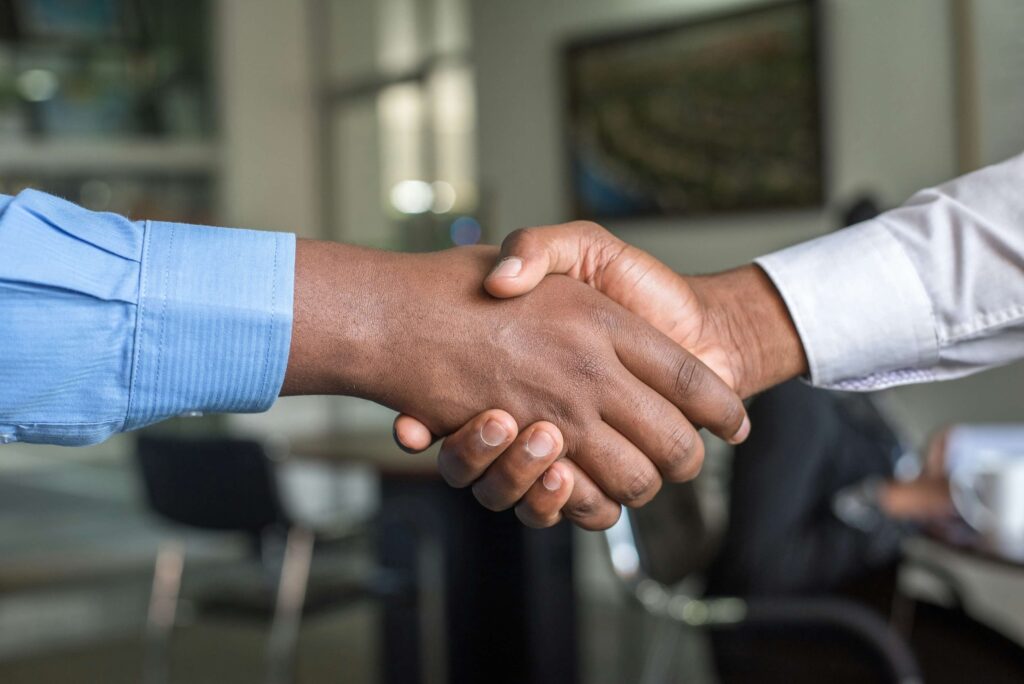 Two people wearing long-sleeved shirts shake hands in an office setting, symbolizing a successful asset lifecycle management strategy, with blurred furniture and a person in the background.