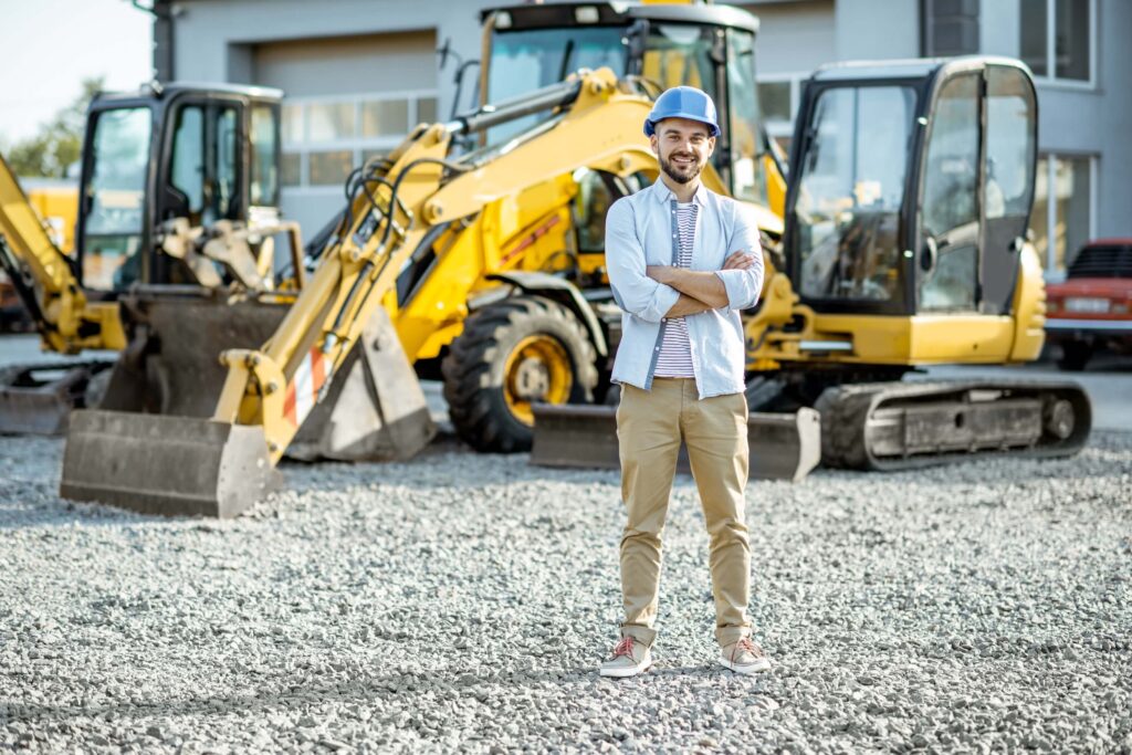 A man wearing a blue hard hat and casual clothes stands with arms crossed on gravel in front of yellow construction vehicles at a work site, embodying the drive needed for anyone learning how to scale a rental business.