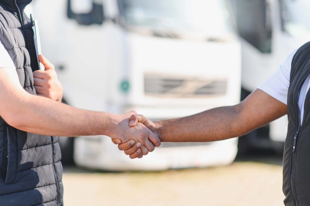 Two people shaking hands outdoors, with white trucks in the blurred background. Only their arms and torsos are visible, suggesting a business agreement or partnership—possibly discussing how to scale a rental business.