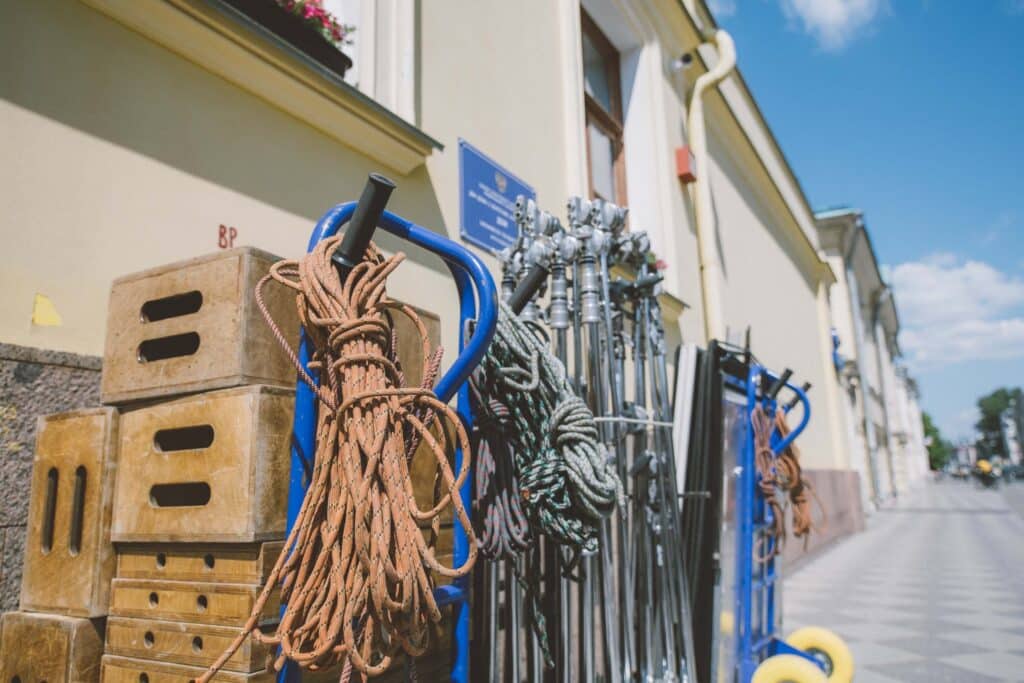 Film production equipment, including wooden boxes, coiled ropes, and metal stands, is stacked on carts outside a beige building under a sunny sky—offering a behind-the-scenes look at how to scale a rental business efficiently.